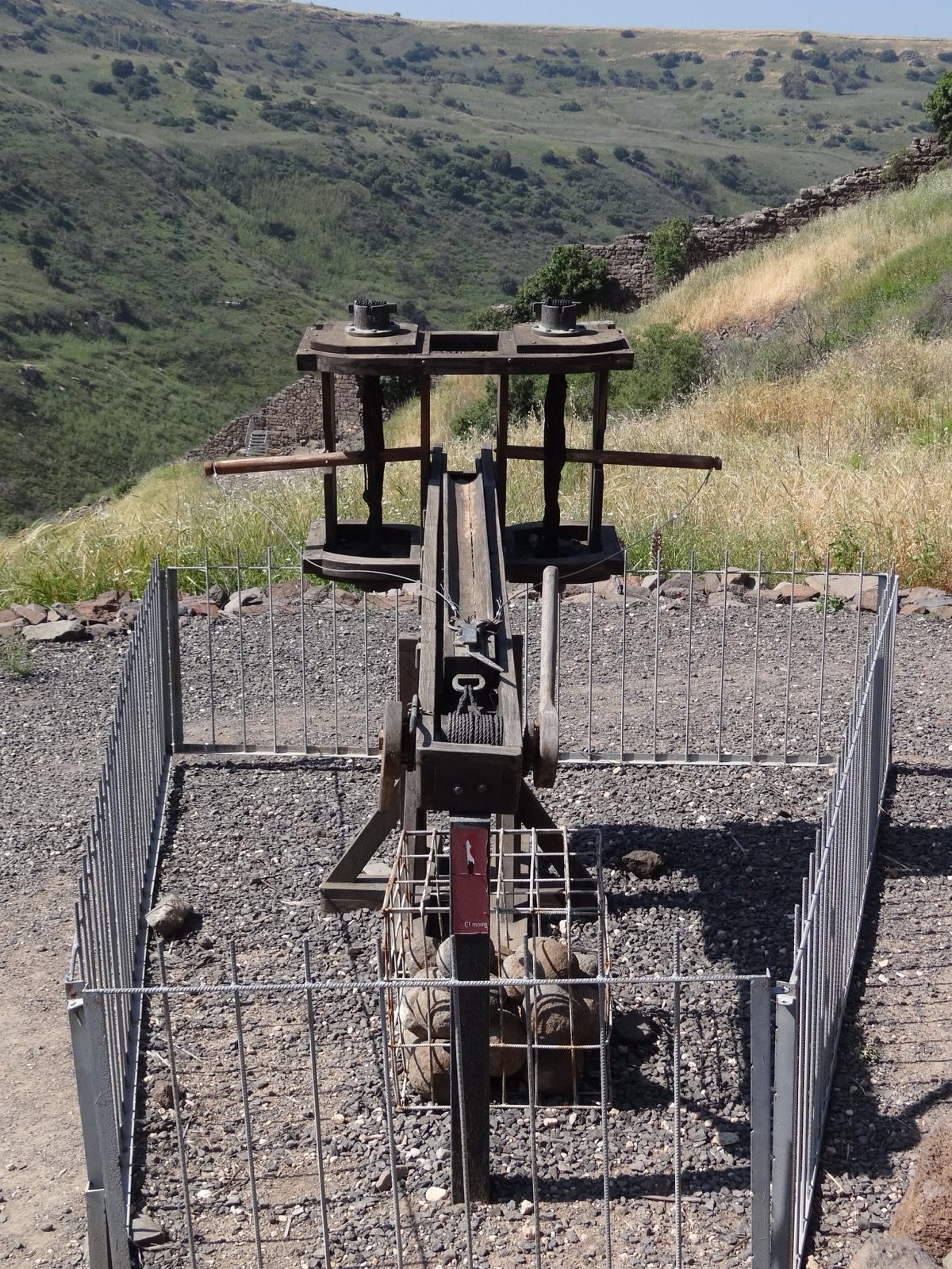 Photograph of a reconstructed Roman ballista model at the Gamla Nature Reserve in Israel, showing its twin torsion arms and frame mounted above a stack of stone ammunition, with the Golan hillside in the background