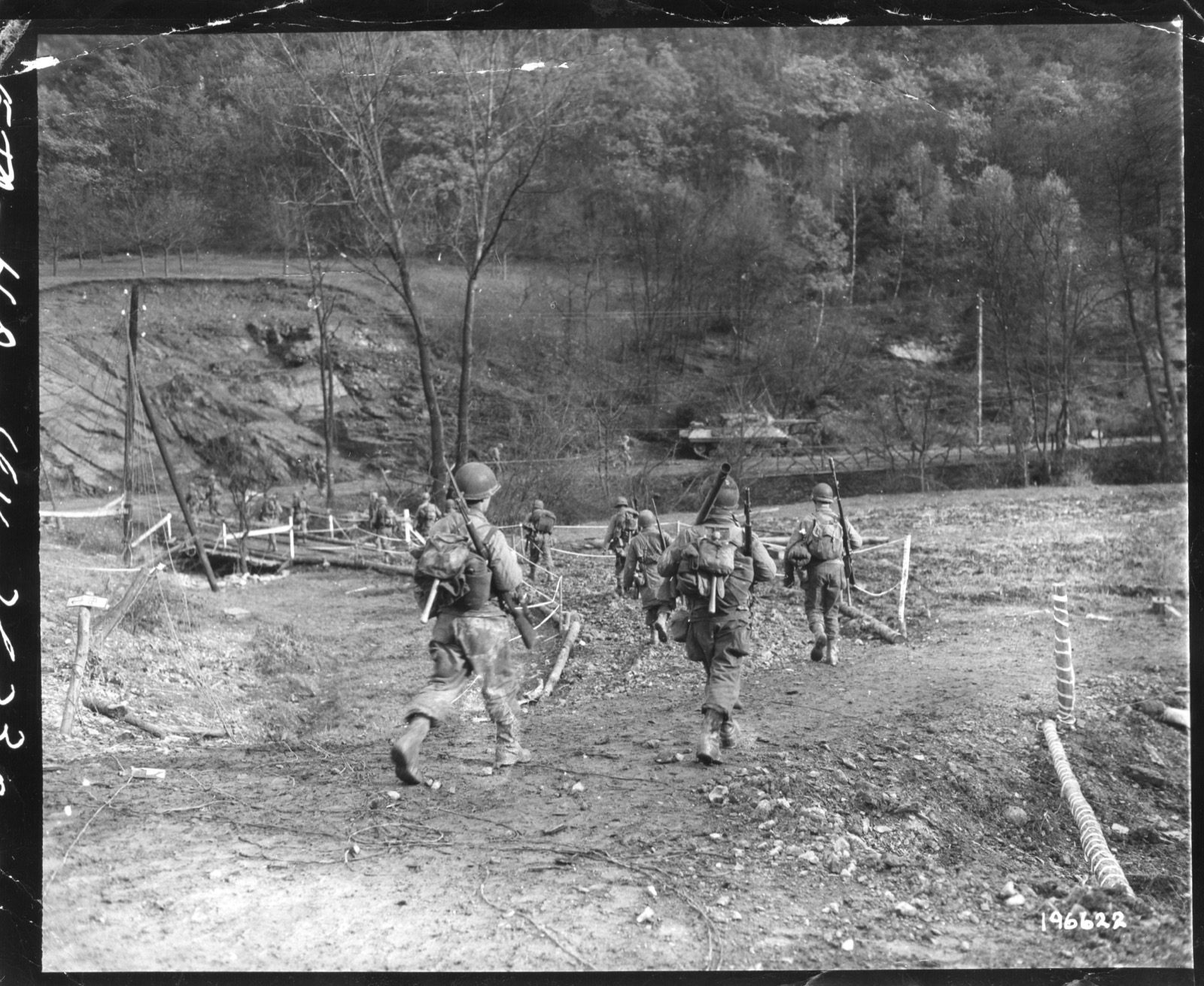 American infantrymen carrying rifles and bazookas cross a log footbridge into the Hürtgen Forest near a Sherman tank, November 18, 1944