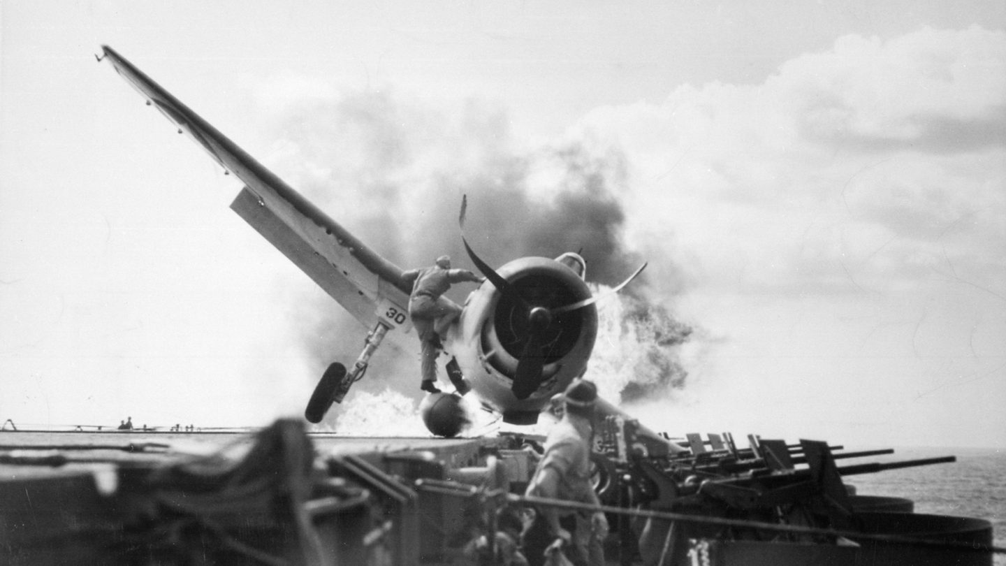 A sailor on a burning Hellcat's wing reaches toward its cockpit while other sailors stand on the carrier deck.