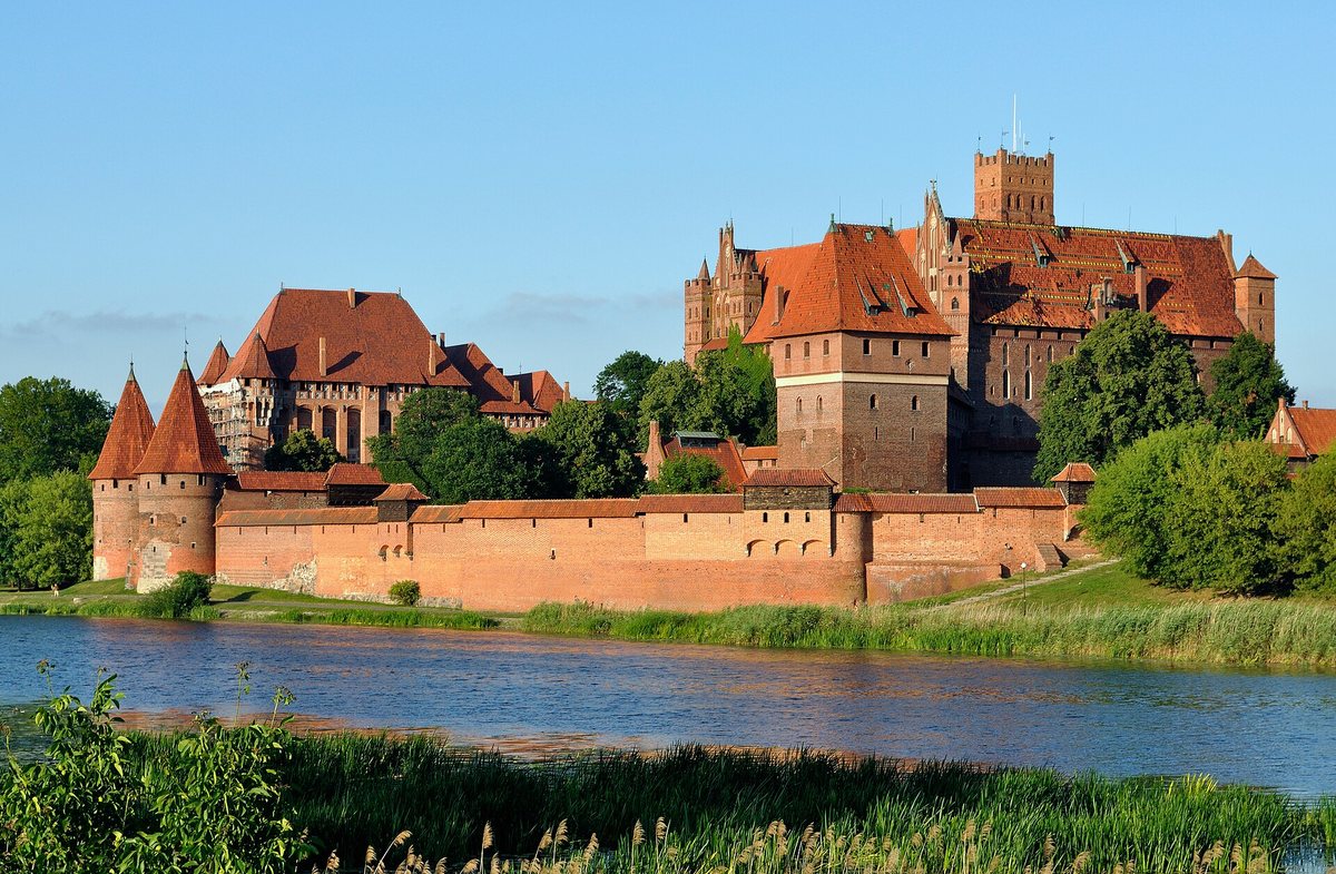 Malbork Castle seen across the River Nogat, a long red-brick fortress with a curtain wall, corner towers and several gabled wings under a clear sky